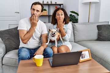 Poster - Hispanic middle age couple sitting on the sofa using computer laptop covering mouth with hand, shocked and afraid for mistake. surprised expression