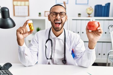 Poster - African american doctor man holding prescription pills and fresh apple angry and mad screaming frustrated and furious, shouting with anger looking up.