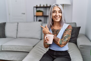 Sticker - Middle age grey-haired woman drinking coffee sitting on the sofa at home cheerful with a smile on face pointing with hand and finger up to the side with happy and natural expression