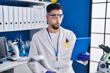 Wall Mural - Young handsome man working at scientist laboratory doing online call scared and amazed with open mouth for surprise, disbelief face