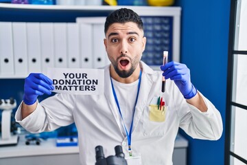 Sticker - Young handsome man working at scientist laboratory holding blood sample afraid and shocked with surprise and amazed expression, fear and excited face.