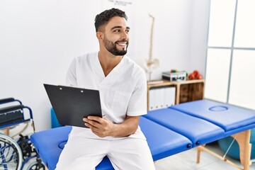 Sticker - Young arab man wearing physiotherapist uniform writing on clipboard at clinic