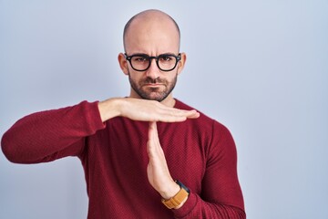 Wall Mural - Young bald man with beard standing over white background wearing glasses doing time out gesture with hands, frustrated and serious face