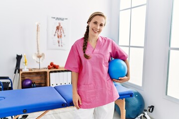 Poster - Young caucasian woman wearing physiotherapist uniform holding fit ball at physiotherapy clinic