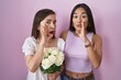 Hispanic mother and daughter holding bouquet of white flowers hand on mouth telling secret rumor, whispering malicious talk conversation