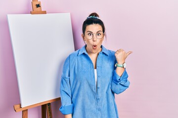 Sticker - Young hispanic woman standing by painter easel stand surprised pointing with hand finger to the side, open mouth amazed expression.