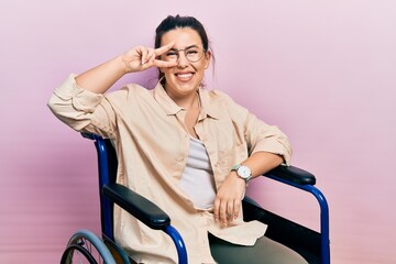 Poster - Young hispanic woman sitting on wheelchair doing peace symbol with fingers over face, smiling cheerful showing victory