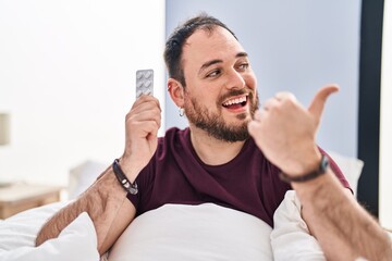 Wall Mural - Plus size hispanic man with beard in the bed holding pills pointing thumb up to the side smiling happy with open mouth