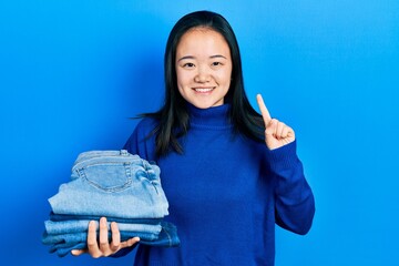 Wall Mural - Young chinese girl holding stack of folded jeans smiling with an idea or question pointing finger with happy face, number one