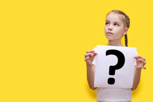 Girl Child Holding A Sheet Of Paper With A Question Mark On A Yellow Background. The Concept Of Curiosity Or Self-doubt. Question Mark On White Paper In The Hands Of A Child. Free Space For Text