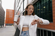 © PhotoBook - Long-haired young european woman is looking on smart watch and holding coffee while going to workplace in office. Active lifestyle of business woman. She is wearing white jacket and shirt