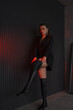 © NASTYA PALEHINA - teen leisure. young girl in black slim dress-jacket is standing fashion and pulling over the knee boots near loft wall background with red studio light. bottom camera. lifestyle concept, free space