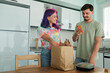 © Evrymmnt - Hipster couple unpacking a paper bag full of take out food containers. Young woman with bright colorful hair and bearded man at the kitchen with delivered food order. Close up, copy space, background