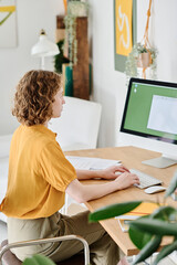 Young freelance webdesigner in casualwear with her hands over keyboard looking at electronic document on computer screen