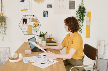 Aufkleber - Side view of young female economist with financial papers using laptop while sitting by workplace and checking or analyzing information