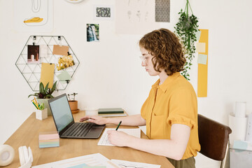 Aufkleber - Young serious businesswoman in casualwear sitting by desk in front of laptop, making notes in financial document and analyzing data