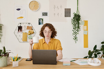 Aufkleber - Young serious freelancer with cup of tea sitting in front of laptop by workplace and searching for online information for project