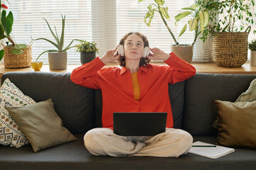 Aufkleber - Young female student in headphones listening to audio lesson while sitting on couch in living room with laptop on her crossed legs
