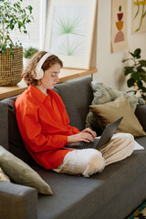 Aufkleber - Youthful female student in red shirt and white pants typing on laptop keyboard while sitting on soft comfortable couch in living room