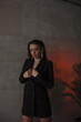 © NASTYA PALEHINA - teen fashion. young girl in black slim jacket is standing fashion on the gray loft wall background with plant in red light and looking with flirt with hands near breast. lifestyle concept, free space