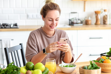 Wall Mural - Healthy lifestyle concept. Beautiful young woman having breakfast and chatting online with friends using mobile phone sitting at home kitchen