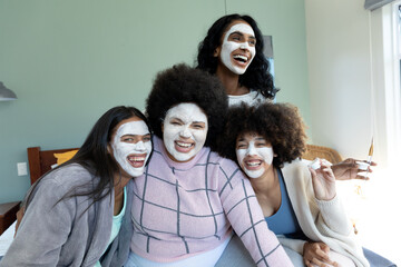 Cheerful biracial millennial female friends with facial cream on faces enjoying leisure time at home