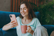 © ViDi Studio - Young happy smiling woman she 20s wear casual clothes mint t-shirt use mobile cell phone hold cup drink coffee tea in morning look camera sit on blue sofa indoor rest at home in own room apartment