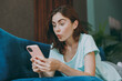© ViDi Studio - Young amazed surprised impressed shocked woman she 20s in casual clothes mint t-shirt hold use mobile cell phone lying on blue sofa indoor rest at home in own room apartment People lifestyle concept.