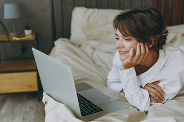 Wall Mural - Young minded fun woman wear white shirt pajama she lying in bed hold use work laptop pc computer study online browsing internet rest relax spend time in bedroom lounge home in own room hotel wake up.