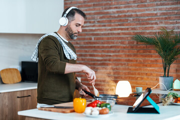 Handsome mature man cooking following a recipe from the internet with digital tablet while listening music with headphones in the kitchen at home.