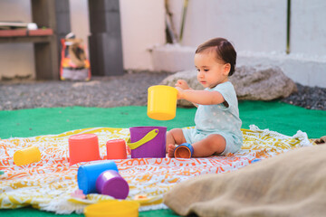 One year old baby playing with colored cubes sitting on a blanket in the garden of his house.