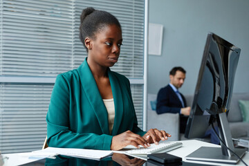 African serious businesswoman typing on keyboard and looking at computer monitor sitting at her workplace at office