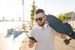 © Julija - Young man holding on his longboard and using a smartphone while walking in the city.
