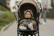 A happy female toddler is holding the safety bumper bar of the stroller on a cloudy day. A young girl in the hood of her raincoat is in her baby carriage in a park at noon.