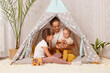 Indoor shot of mother with two kids sitting together in wigwam teepee at home, mom playing with preschool and infant children, showing wooden eco toy to her baby.