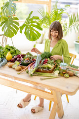 Wall Mural - Portrait of a young cheerful woman sits by the table full of fresh vegetables, fruits and greens indoors with plants on background. Healthy eating and lifestyle concept