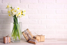Daffodils In A Glass Vase With Gifts Against A White Brick Wall. Copy Space.