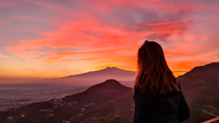 Wall Mural - Tourist woman watching beautiful sunset behind volcano Mount Etna near Castelmola, Taormina, Sicily, Italy, Europe, EU. Clouds with vibrant red orange colors. Silhouette of person during twilight