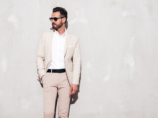 Portrait of handsome confident stylish hipster lambersexual model. Sexy modern man dressed in white elegant suit. Fashion male posing in studio near grey wall in sunglasses