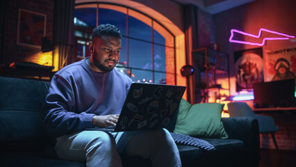 Fototapete - Young Stylish Black Man Working from Home on Laptop Computer with Stickers in Cozy Stylish Loft Apartment. Creative Male Smiling, Checking Social Media, Browsing Internet. Flat with Big Windows.