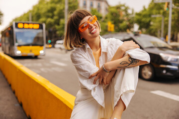 Wall Mural - Cool young caucasian woman in good mood looking away posing outside. Brown-haired wears orange glasses, white casual clothes. City life concept