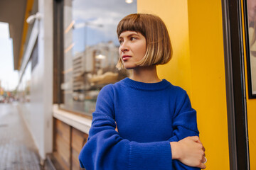 Wall Mural - Serious young caucasian girl looks away, arms crossed standing outdoors. Brown-haired woman with bob haircut wears blue sweatshirt in spring. City life concept