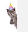 Winking cat wearing birthday cap holds microphone above empty white banner. isolated on white background