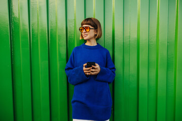Wall Mural - Cute young caucasian girl with phone in her hands looks to side standing against green wall. Brown-haired woman wears sunglasses, blue sweater autumn. City life concept