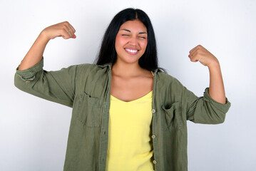 Strong powerful young brunette woman wearing green overshirt standing against white background toothy smile, raises arms and shows biceps. Look at my muscles!