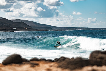 Wild rocky coastline of surf spot La Santa Lanzarote, Canary Islands, Spain. Surfer riding a big wave in rocky bay, volcano mountain in background.