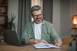 © Prostock-studio - Smiling elderly european male with beard working with laptop, watch video lesson or webinar in living room