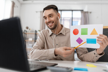 Wall Mural - distance education, primary school and remote job concept - happy smiling male teacher with laptop and picture of geometric shapes in different colors having online class at home office