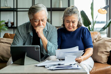 Old Retired Asian Senior Couple Checking And Calculate Financial Billing Together On Sofa Involved In Financial Paperwork, Paying Taxes Online Using E-banking Laptop At Living Room Home Background