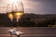 Two elegant glasses of white wine on the right, and a countryside view in the background, at sunset